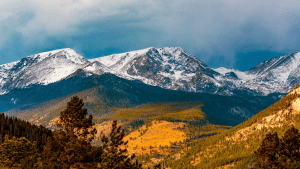 Image of the Colorado Rocky Mountains in the fall. In the foreground is valleys of pine trees and autumn grass.